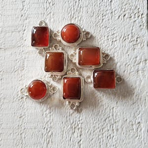 Può includere: Sette componenti di gioielleria argentati con pietre preziose rosso-arancio lucide in varie forme: rotonde, quadrate e rettangolari. Ogni pezzo ha una cornice decorativa argentata e piccoli anelli per il fissaggio. I componenti sono disposti su una superficie bianca strutturata.