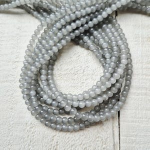 May include: Multiple strands of grey gemstone beads. The beads are small, round, and have a smooth, polished surface. The beads are strung together, forming multiple strands. The colour is a soft, muted grey, with a slight translucence. The beads are arranged in a circular pattern.