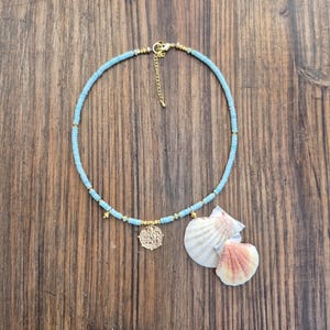 May include: A necklace featuring light blue disc-shaped beads interspersed with small gold-toned metallic spacers. The piece includes a circular gold-toned coin pendant and two natural white and pinkish seashells attached as charms, with a gold-toned clasp.
