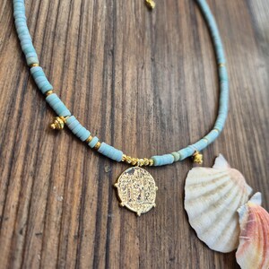 May include: A necklace featuring light blue disc-shaped beads interspersed with small gold-toned metallic beads. The piece includes a central gold-toned circular pendant with an embossed design and two smaller gold-toned charms, finished with a gold-toned chain extender.