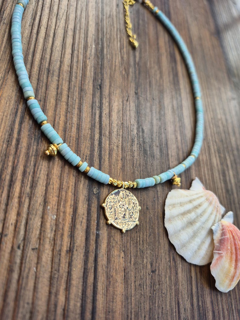 May include: A necklace featuring light blue disc-shaped beads interspersed with small gold-toned metallic beads. The piece includes a central gold-toned circular pendant with an embossed design and two smaller gold-toned charms, finished with a gold-toned chain extender.