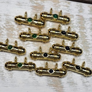 May include: Ten gold-colored metal charms with green and black gemstones. Each charm is shaped like a cylinder with a decorative design. The charms are arranged in a random pattern on a light wood surface.