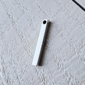 May include: A small, rectangular, silver pendant with a hole at the top. The pendant is lying on a light grey, textured surface. The pendant has a simple, minimalist design.