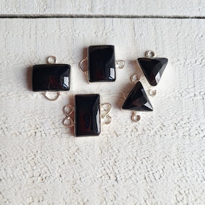 May include: Five black gemstone pendants in various shapes: square, rectangular, and triangular. Each pendant has a silver-colored bail for attaching to a chain or bracelet. The gemstones are polished and reflect light.