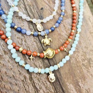 May include: Multiple beaded necklaces with gold charms. The necklaces feature blue, white, and orange beads. Charms include a turtle, a cross, and a shell. The necklaces are displayed on a wooden surface.