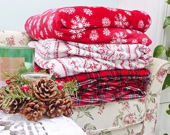 May include: A stack of three folded blankets in red and white holiday patterns. The top blanket is red with white snowflakes, the middle one is white with red and gray patterns, and the bottom one is red plaid.