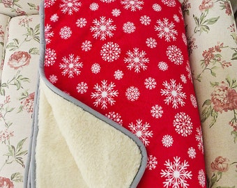 May include: A red fleece blanket with a white snowflake pattern. The blanket is folded in half and has a cream-colored fleece lining.