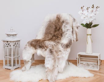 May include: A white faux fur rug with a brown and white reindeer hide rug draped over a chair. The chair is in front of a white wall and there is a white vase with white flowers on a white wooden crate.