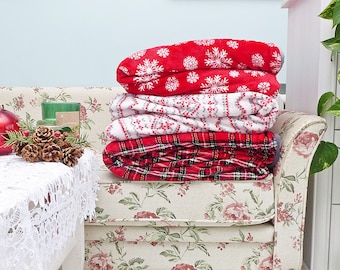 May include: A stack of four red and white patterned fleece blankets on a floral patterned couch. The blankets feature snowflake, plaid, and geometric designs.