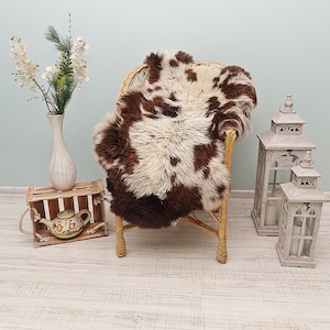 May include: A brown and white spotted sheepskin rug draped over a light brown wicker chair. To the left, a wooden crate holds a ceramic teapot and a white vase with white flowers. To the right, two white wooden lanterns stand on the floor.