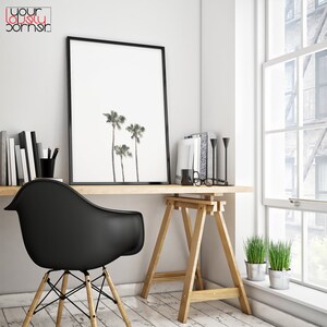 May include: A black framed print of three palm trees against a white background hangs above a wooden desk with a black chair. The desk is supported by two wooden sawhorses. A potted plant sits on the floor next to the desk.