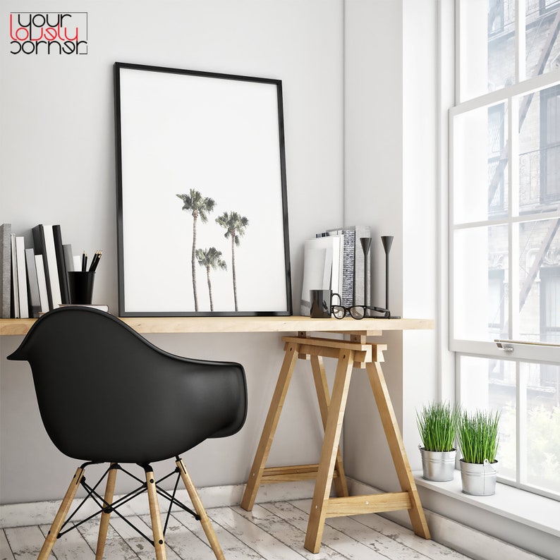 May include: A black framed print of three palm trees against a white background hangs above a wooden desk with a black chair. The desk is supported by two wooden sawhorses. A potted plant sits on the floor next to the desk.