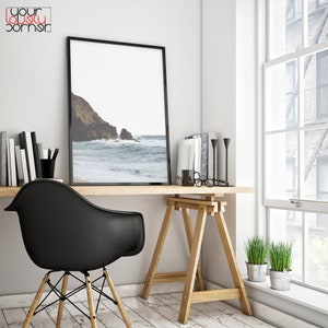 May include: A black framed photograph of a beach scene with waves crashing on the shore. The photo is hanging on a white wall above a wooden desk with a black chair and a potted plant.