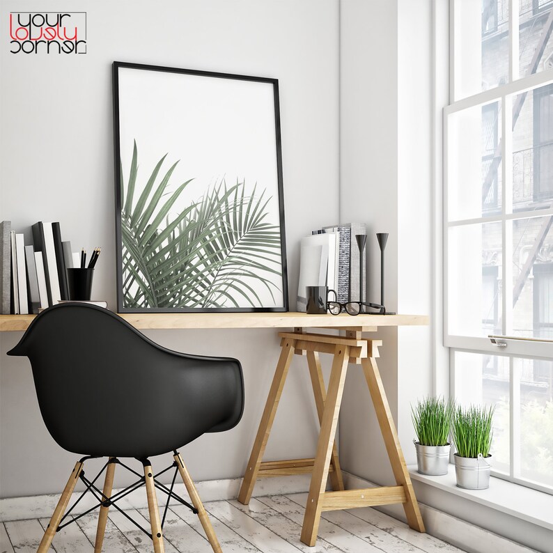 May include: A black framed print of a green palm leaf against a white background hangs above a wooden desk with a black chair. The desk is supported by two wooden sawhorses. There are two small potted plants on the floor next to the desk.