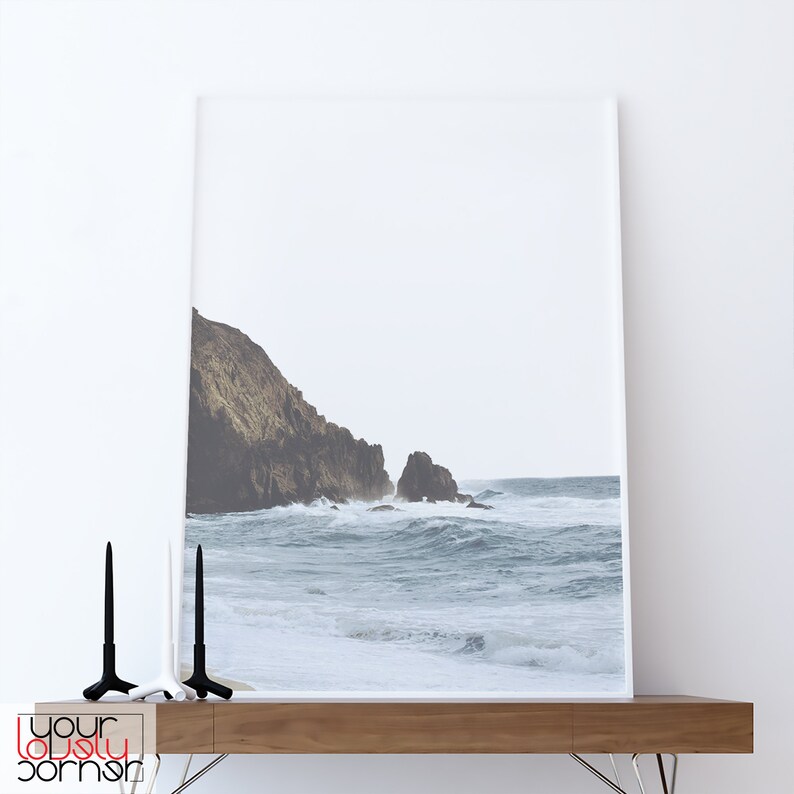 May include: A framed photograph of a rocky coastline with crashing waves. The photo is in a white frame and is hanging on a white wall.