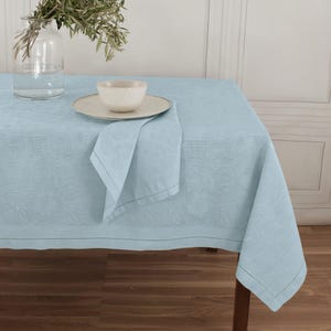 Peut inclure: Une nappe bleu clair avec un motif floral subtil, une serviette assortie, un bol et une assiette en céramique beige. La nappe est posée sur une table en bois, créant un cadre serein et élégant. Un vase en verre transparent avec des tiges vertes est en arrière-plan.