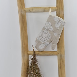Peut inclure: Un gant de cuisine beige avec un motif floral blanc est suspendu à une échelle en bois. Le gant présente un motif d'hortensia et une bordure crème. Un bouquet d'herbes séchées est attaché à l'échelle en dessous.