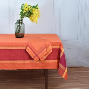 May include: An orange tablecloth with a decorative border and matching napkins. The tablecloth is set on a dark brown table. A glass vase with yellow flowers sits on the table, adding a touch of colour.