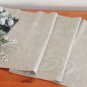 May include: A beige paisley patterned table runner is displayed on a wooden surface. A black and white floral tea set sits on a matching tray. A small bowl of white cookies and white flowers complete the scene.