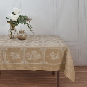 Könnte beinhalten: Eine beige rechteckige Tischdecke mit floralem Muster bedeckt einen Holztisch. Zwei Glasvasen mit weißen Blumen und Grün stehen auf dem Tisch. Die Tischdecke hat ein sich wiederholendes Design mit floralen Motiven.
