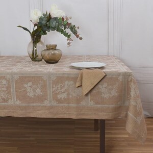 Può includere: Una tovaglia beige con un motivo floreale, appoggiata su un tavolo di legno. Un piatto bianco e un tovagliolo beige piegato sono sul tavolo. Un vaso con fiori bianchi e verde, e un vaso marrone sono anche sul tavolo.