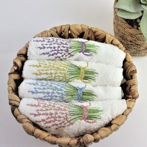 以下が含まれることがあります： 織りバスケットに入った、刺繍されたラベンダーの小枝が付いた、4つの白い丸められたタオル。各タオルは、紫、黄色、青、ピンクの異なる色のラベンダーが特徴です。タオルは丸い茶色の籐のバスケットにきちんと配置されています。