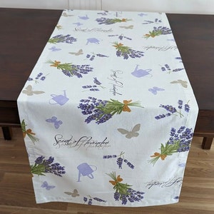 Può includere: Runner da tavolo bianco con un design alla lavanda. Presenta mazzi di lavanda viola legati con spago marrone, annaffiatoi alla lavanda e farfalle beige. Il testo "Scent of Lavender" è stampato sul tessuto.