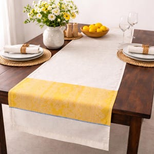 May include: A white and yellow table runner with a floral design, set on a dark wooden table. The runner has a yellow band with a detailed pattern and a thin blue stripe. The table is set with plates, glasses, and a vase of flowers.