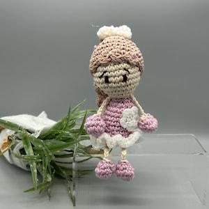 May include: A handmade crochet doll in a pink dress with white trim and a matching hat. The doll has a light brown complexion, a white flower accent, and small pink pom-poms for hands and feet. The doll is approximately 10 cm tall.
