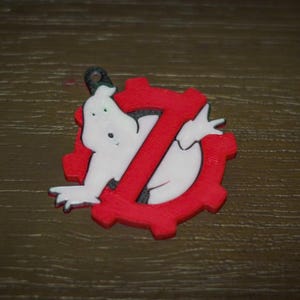 May include: A white cartoon ghost figure emerging from a red gear-shaped design with a red "Z" in the center. The design has a black loop at the top for hanging. The overall theme is related to the Ghostbusters franchise.