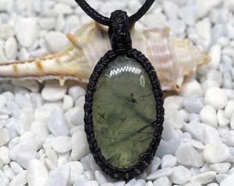 May include: A green aventurine gemstone pendant with black cord necklace. The pendant is oval shaped and has a dark green color with black and white inclusions. The necklace is made of black cord and is tied with a knot at the top.