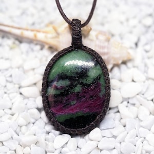 Könnte beinhalten: Ein grüner, rosa und schwarzer Edelstein-Anhänger an einer braunen Schnurkette. Der Anhänger ist oval geformt und in einem braunen Makramee-Knoten gefasst.