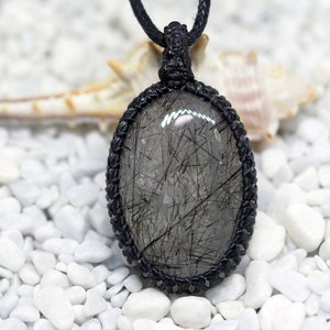 Pu&ograve; includere: Una collana con cordoncino nero con un ciondolo ovale argentato e nero che presenta un cristallo di quarzo trasparente con inclusiones nere. La collana &egrave; esposta su un letto di ciottoli bianchi.