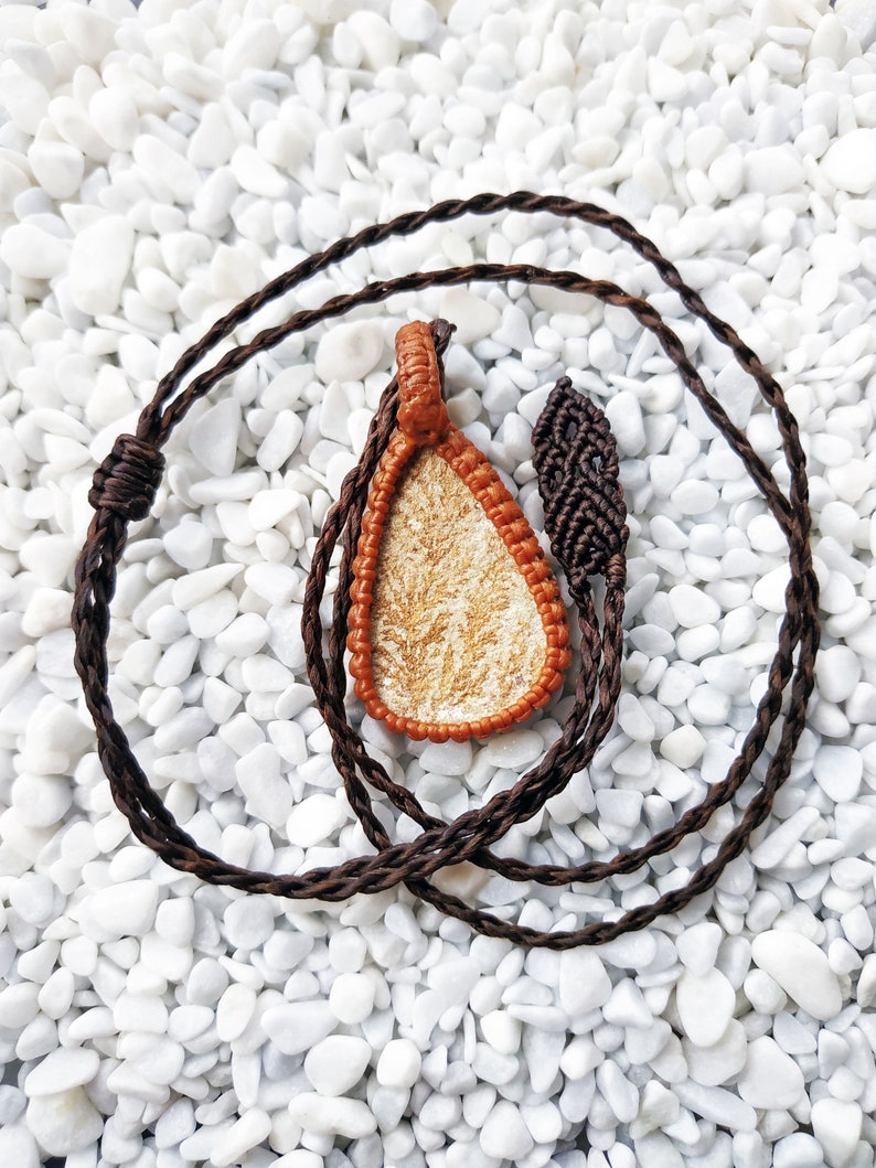 May include: A brown cord necklace with a teardrop-shaped pendant. The pendant is made of a light brown material with a white pattern. The necklace is lying on a bed of white pebbles.