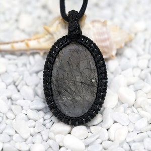 Pu&ograve; includere: Una collana con cordoncino nero con un ciondolo ovale argentato e nero con un cristallo di quarzo trasparente con inclusioni nere. Il ciondolo &egrave; su uno sfondo bianco di piccole pietre bianche.