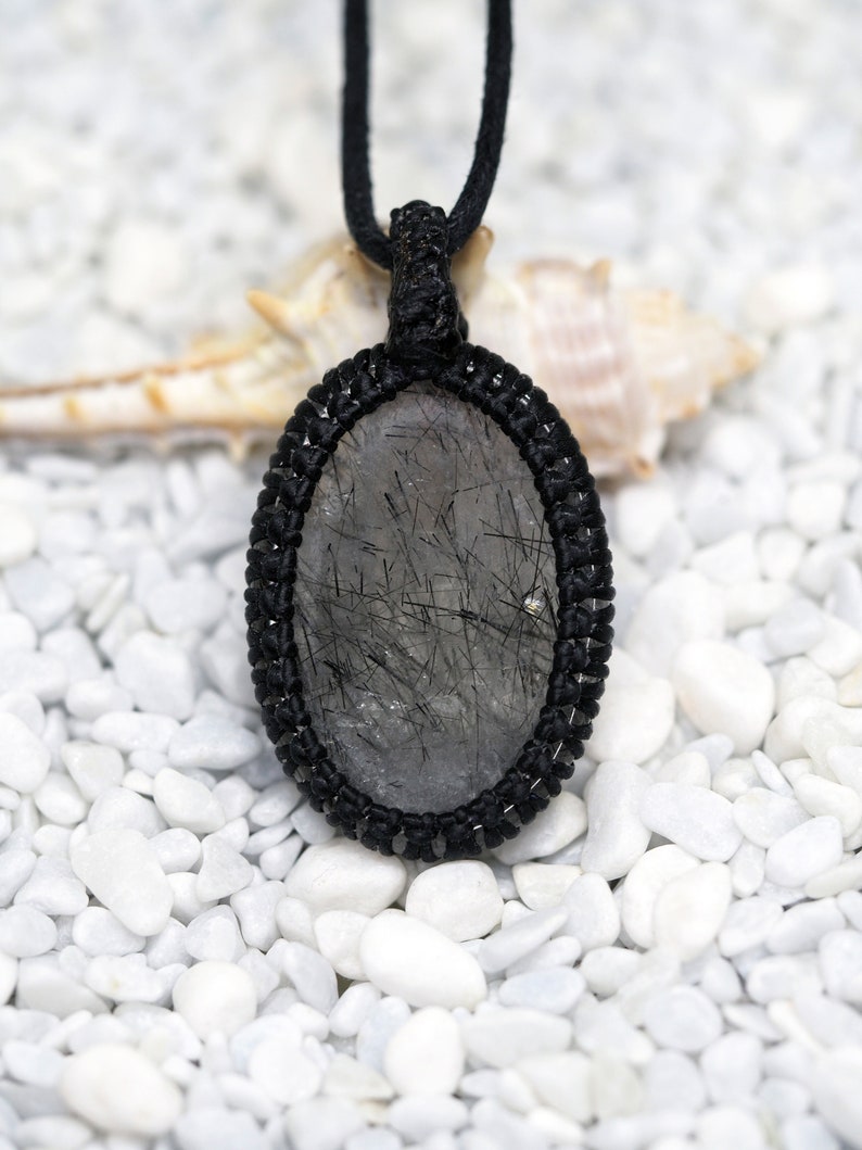 Pu&ograve; includere: Una collana con cordoncino nero con un ciondolo ovale argentato e nero con un cristallo di quarzo trasparente con inclusioni nere. Il ciondolo &egrave; su uno sfondo bianco di piccole pietre bianche.