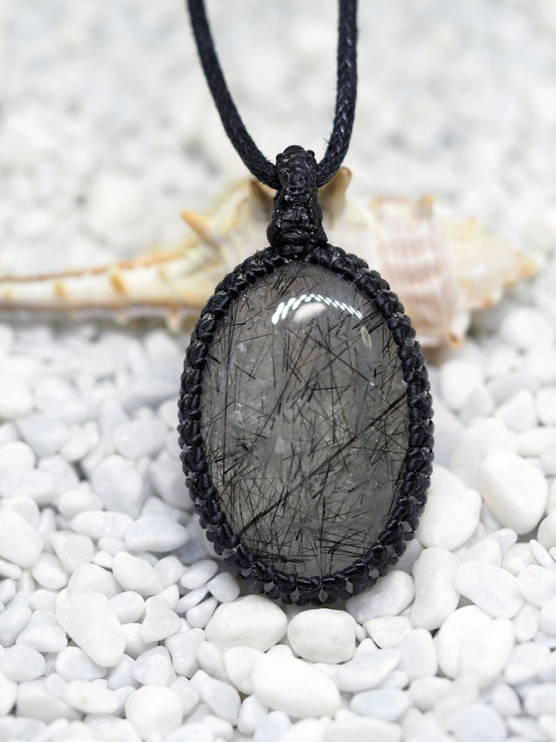 Pu&ograve; includere: Una collana con cordoncino nero con un ciondolo ovale argentato e nero che presenta un cristallo di quarzo trasparente con inclusiones nere. La collana &egrave; esposta su un letto di ciottoli bianchi.