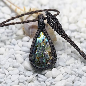 Puede incluir: Un collar de cordón marrón con un colgante de concha de abulón en forma de lágrima. El colgante tiene colores iridiscentes verdes, azules y morados. El collar está sobre un fondo blanco.