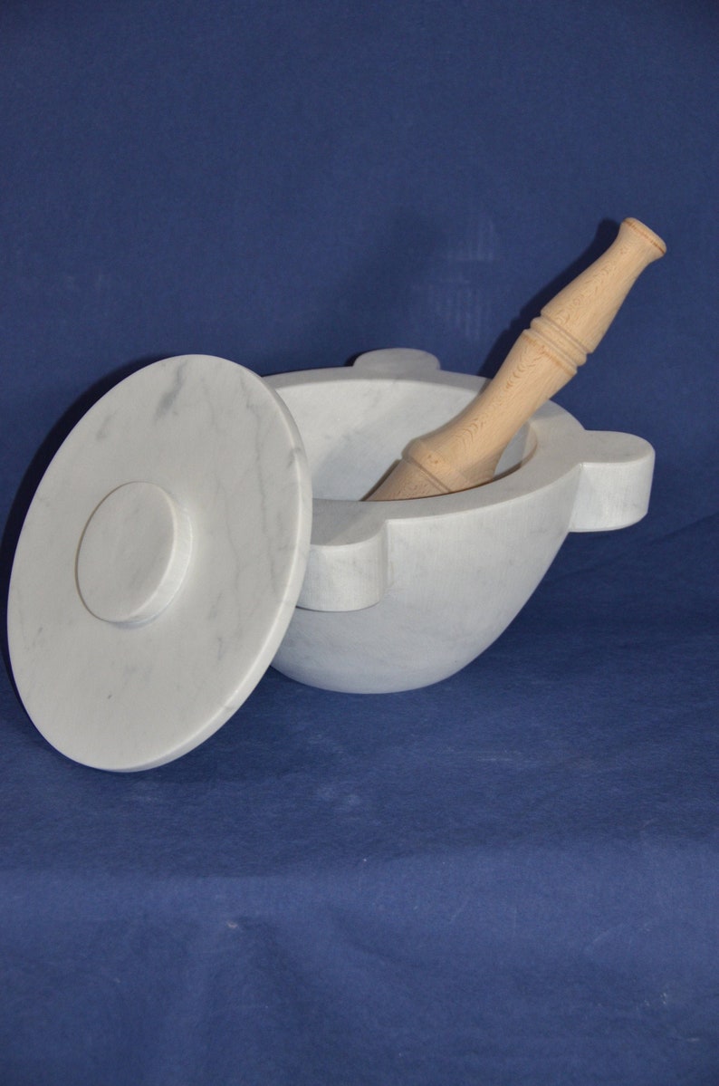 Może przedstawiać: Moździerz i tłuczek z białego marmuru z drewnianym tłuczkiem. Moździerz ma pokrywkę i zaokrąglone dno. Tłuczek jest gładki i ma zaokrąglony koniec.