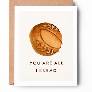 May include: A card with a watercolor illustration of a brown bread roll with a bite taken out of it. The text on the card reads "You are all I knead."