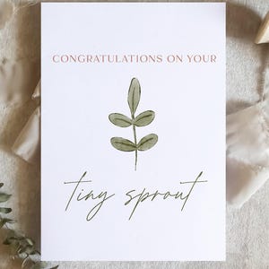 May include: A white card with the words "CONGRATULATIONS ON YOUR tiny sprout" in a script font. The card has a green leaf illustration. A beige ribbon and eucalyptus sprig are arranged around the card on a neutral background.