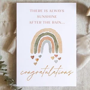 May include: A greeting card with a watercolor rainbow design in muted tones. The card reads "THERE IS ALWAYS SUNSHINE AFTER THE RAIN... congratulations" in elegant script. Small heart illustrations are below the rainbow.