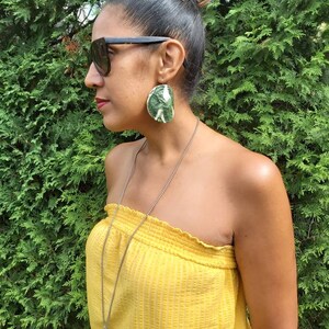 Può includere: Una donna indossa un top giallo senza spalline e occhiali da sole neri con grandi orecchini a forma di foglie verdi e bianche.