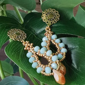 May include: A pair of gold and light blue earrings with a delicate filigree design. The earrings feature a teardrop shape with multiple layers of beads.