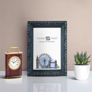 May include: A black picture frame with an ornate design. The frame is empty and has a white mount. The text "TAILORED FRAMES' THE FINEST PICTURE FRAMES" is printed on the mount.