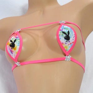 May include: A neon pink bikini set with a tie-dye pattern and the Playboy bunny logo. The bikini top and bottom are connected by thin pink straps and embellished with small, sparkling accents. The set is displayed on a mannequin.