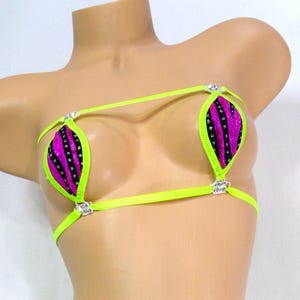 May include: A neon green and purple bikini set. The bikini top features two triangle cups with a purple and black patterned design, outlined in neon green. The bottoms are a matching thong style. Rhinestone accents are placed on the straps.