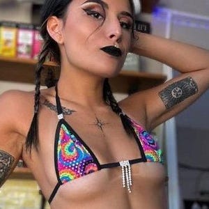 May include: A woman wearing a colorful tie-dye bikini top with black straps and a matching tie-dye bikini bottom with black straps. The bikini top has a silver chain with rhinestones hanging from the center. The woman has black hair in braids and is wearing black eyeliner and lipstick.