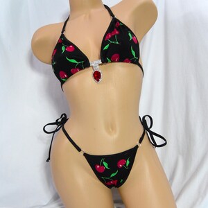 May include: Black bikini set with a cherry print. The triangle top features a rhinestone and red gem embellishment. The bikini bottom has tie sides. The cherries are red with green leaves.