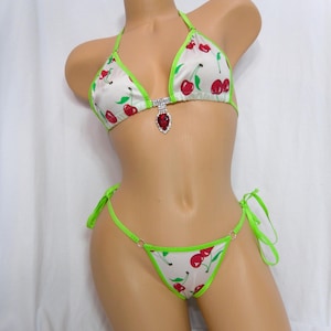 May include: White bikini set with a cherry print. The bikini top has a halter neck and a jewel accent. The edges are trimmed in neon green, and the bottoms are a string style.
